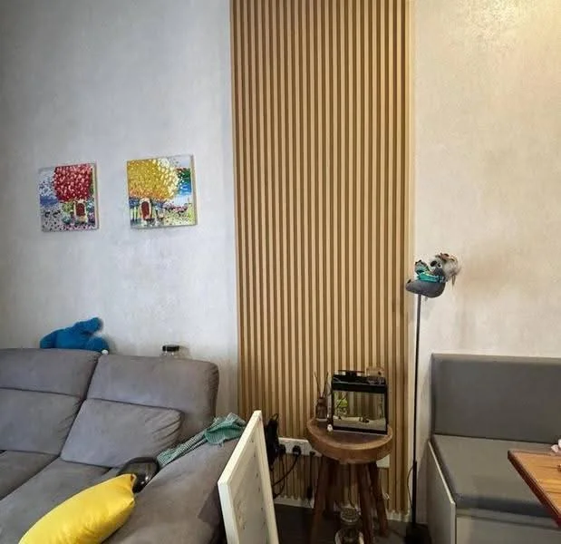 Living features: grey fabric sofa, wooden side table, decorative wall art with colorful trees, textured wall paneling in light wood, light-colored painted walls, small decorative items on the side table, yellow throw pillow, corner seating area, flooring not visible; style: modern.