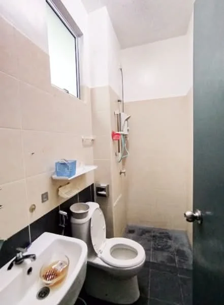 Bathroom features: white ceramic toilet, single-sink vanity with wall-mounted faucet, shower area with wall-mounted showerhead, grey tiled flooring, beige tiled walls, mirror above sink, toilet paper holder, small shelf above toilet, window for natural light.
