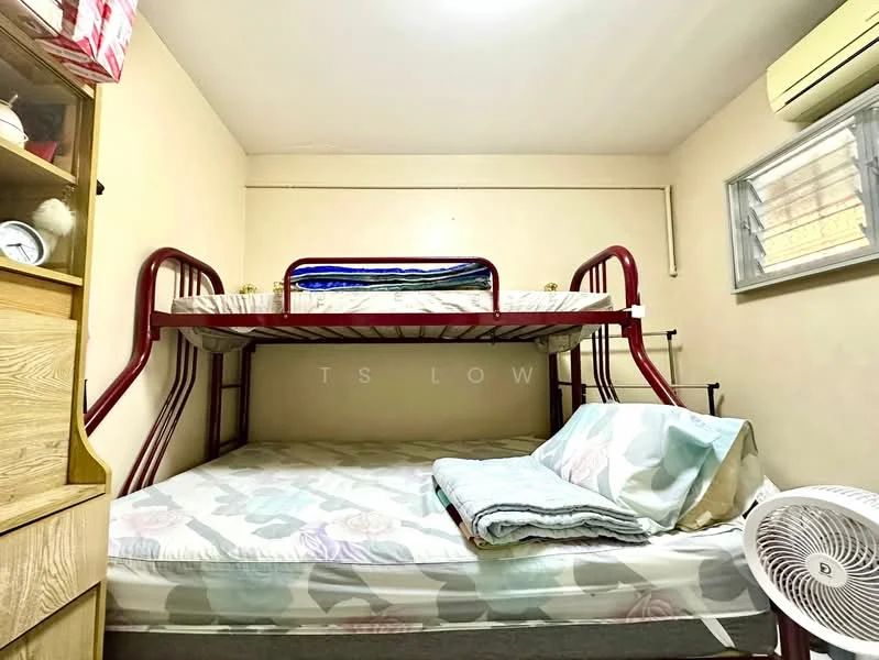 Bedroom features: bunk bed with red metal frame, mattress on top bunk, mattress on bottom bunk, light-colored walls, window with blinds, wall-mounted air conditioning unit, fan on the floor, wooden shelving unit, decorative items on shelf.