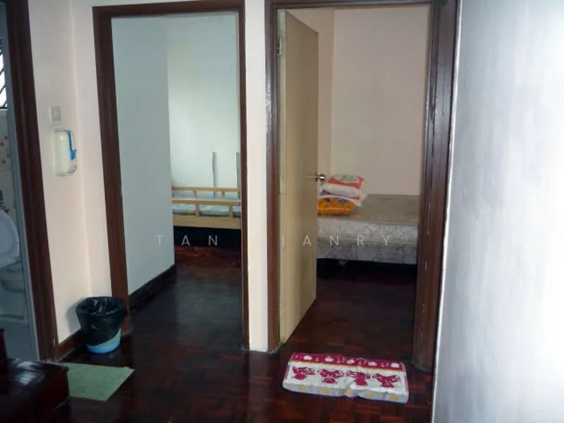 Room features: wooden flooring, white painted walls, brown wooden door, light-colored bedding on bed, bed frame, wastebasket, doorway leading to another room, rug with floral pattern.