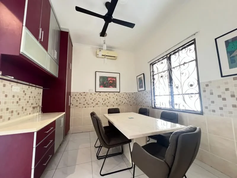 Dining features: white walls, cream tiled flooring, ceiling fan, wall-mounted split AC, rectangular dining table, 6 upholstered dining chairs, red cabinetry with glass doors, cream countertop, decorative wall art, window with decorative iron grille; style: modern.