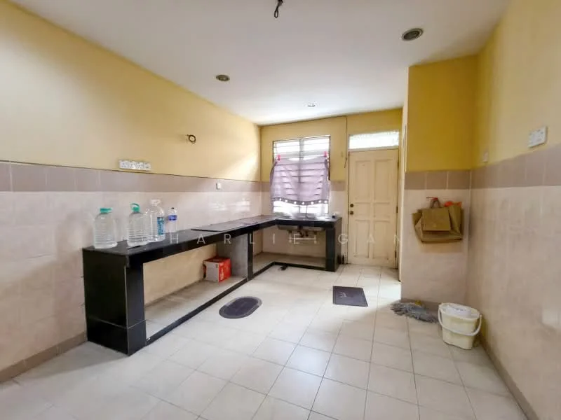 Kitchen features: black countertop, cream tiled walls, light yellow painted walls, floor tiles, single-bowl stainless-steel sink, window with sheer curtains, wooden door, plastic bucket, bottled water containers, wall-mounted hooks.