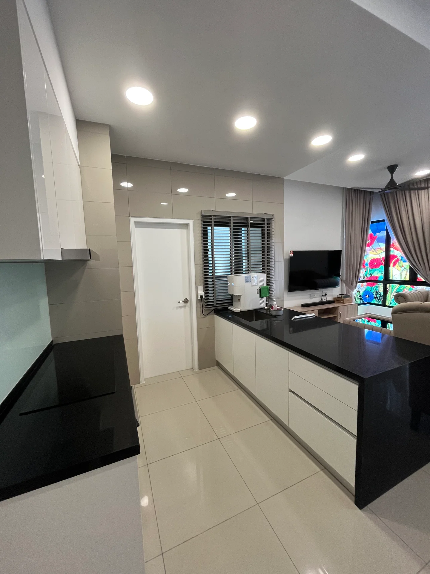 Kitchen features: white cabinetry, black countertop, ceramic tile flooring, recessed downlights, single-bowl stainless-steel sink, chrome pull-down faucet, built-in microwave, window with blinds, open layout to living area, wall-mounted TV, decorative plants, light-colored walls, island counter with seating; style: modern; view: garden.