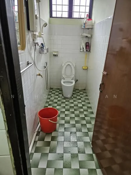 Bathroom features: white tiled walls, green and white checkered tile flooring, one-piece toilet, shower head with wall-mounted faucet, mirror above sink, shelf for toiletries, window for natural light, bucket for water.