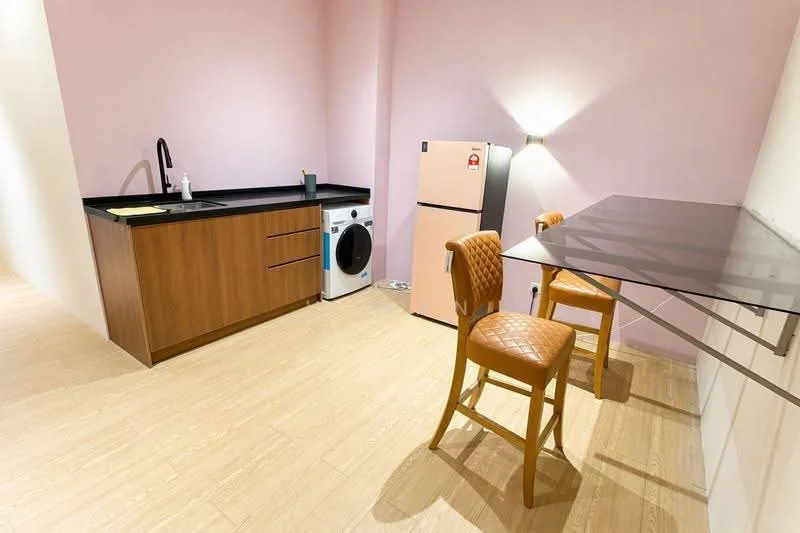 Kitchen features: pink painted walls, black countertop, wood cabinetry, single-bowl stainless-steel sink, chrome pull-down faucet, white washing machine, stainless-steel fridge, glass dining table, 2 upholstered dining chairs, laminate flooring; style: modern.