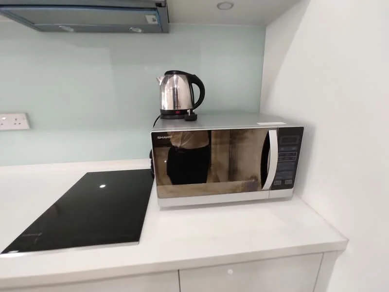 Kitchen features: microwave, stainless-steel kettle, black glass cooktop, white countertop, light green backsplash, under-cabinet lighting, wall-mounted cabinets, electrical outlets; style: modern.