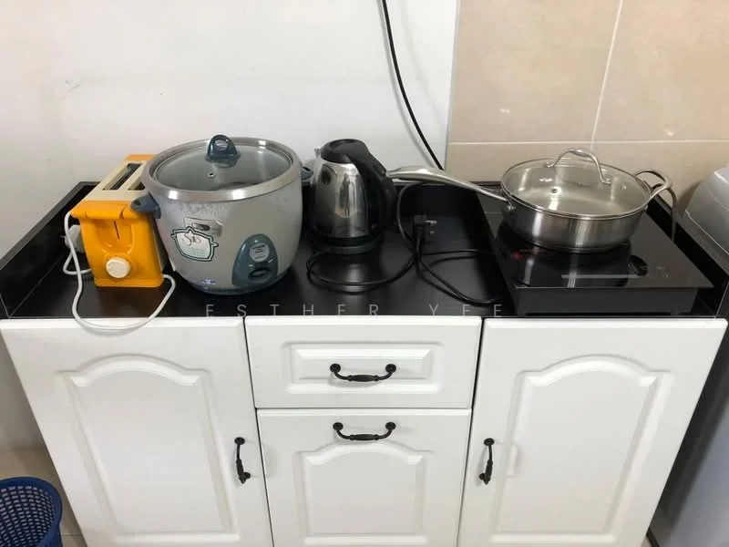 Kitchen features: black countertop, white cabinetry with decorative handles, electric stove, stainless-steel pot with lid, electric kettle, rice cooker, toaster, wall-mounted power outlet.