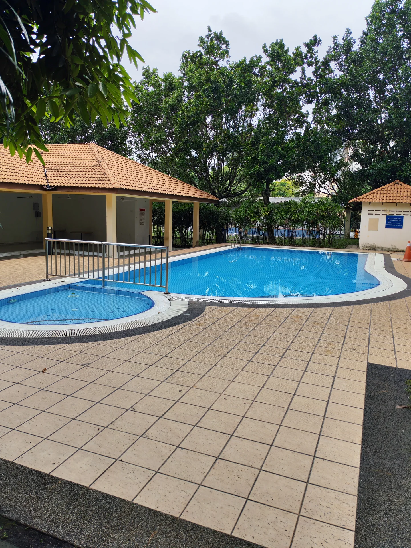 Exterior type: swimming pool deck; features: rectangular swimming pool, jacuzzi area, tiled pool deck, surrounding greenery, covered pavilion with tiled roof, fenced pool area, poolside seating area, landscaped garden, outdoor lighting, non-slip flooring; style: tropical-modern.