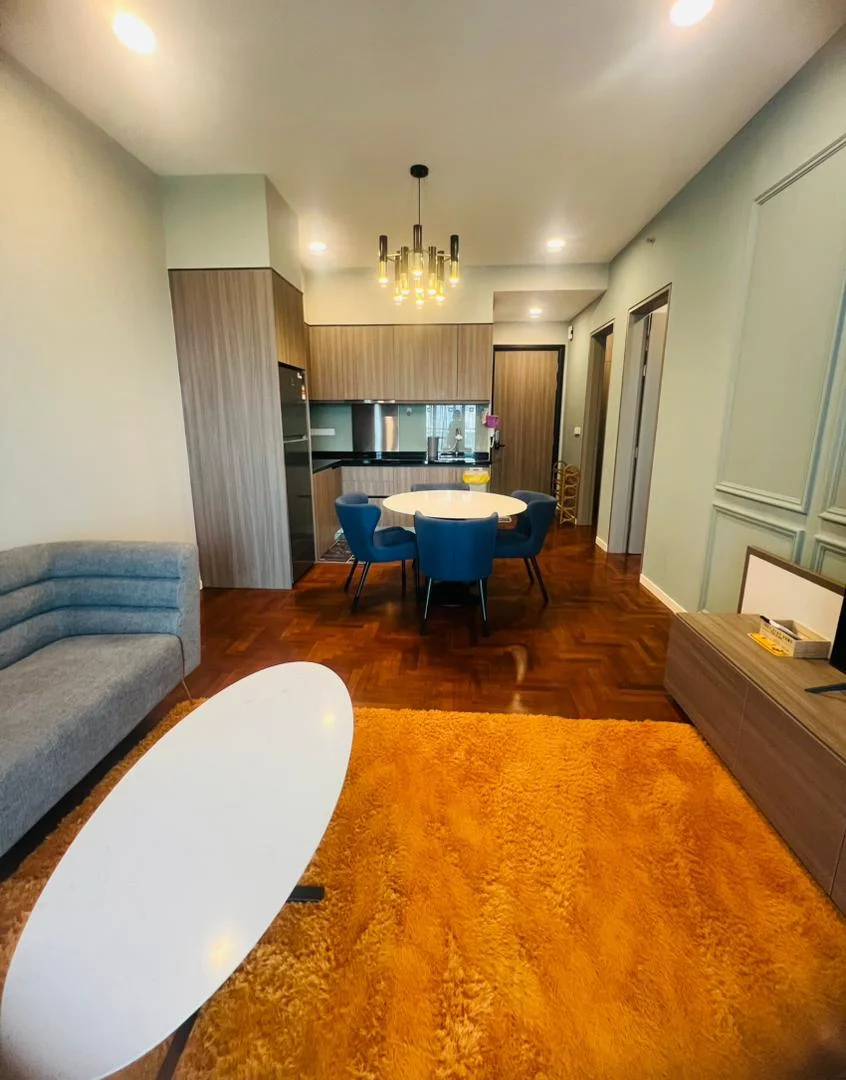 Living features: grey fabric sofa, round dining table, 4 upholstered dining chairs in blue, chandelier with multiple bulbs, wooden flooring, area rug in orange, wall-mounted TV cabinet, decorative wall paneling, recessed ceiling lights, open kitchen layout; style: modern.