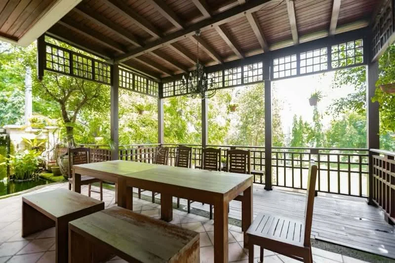 Balcony features: wooden dining table, wooden benches, wooden ceiling, exposed beams, screened windows, potted plants, tile flooring, greenery view; style: traditional; view: garden.