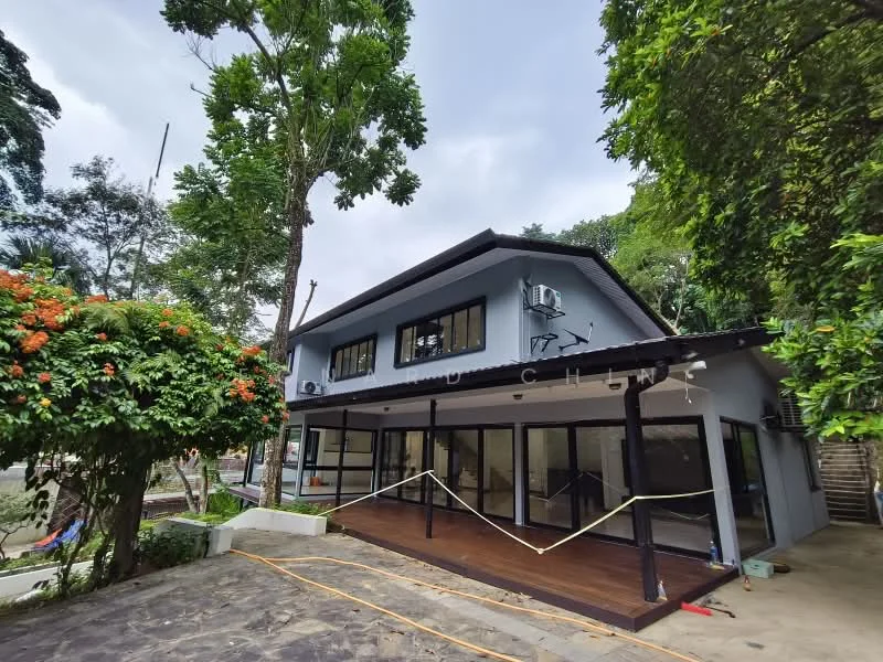 Exterior type: detached bungalow; features: grey painted facade, flat roof, large glass windows, wooden deck, air conditioning units visible, manicured landscaping, tropical trees surrounding the property, stone-paved walkway, outdoor seating area, fenced perimeter; style: modern.
