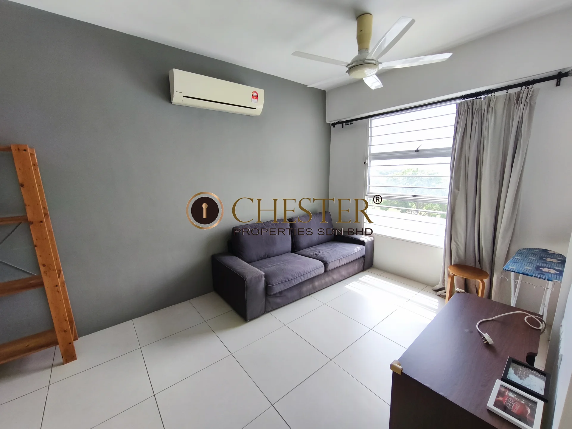 Living features: grey accent wall, white painted walls, white ceiling, 5-blade ceiling fan with light, large window with sheer curtains, sliding window, tile flooring, sofa in dark grey, wooden bookshelf, wooden desk, air conditioning unit on wall; style: modern; view: garden.