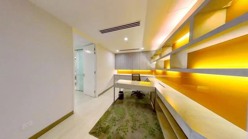 Study features: light wood flooring, cream painted walls, built-in shelving with backlighting, desk with light wood finish, black office chair, green area rug with pattern, recessed ceiling lights, glass door leading to another room; style: modern.