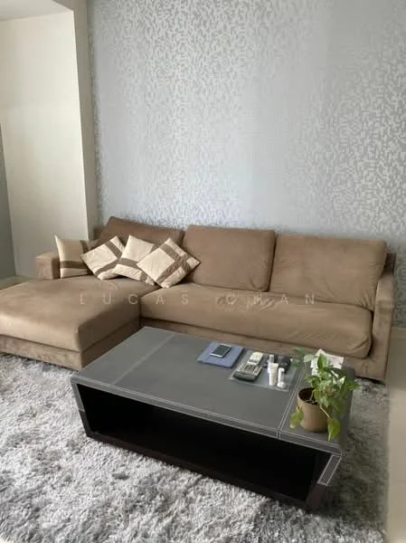 Living features: L-shape sectional sofa in beige, coffee table with grey top, area rug with plush texture, decorative cushions with geometric pattern, wall with textured wallpaper, plant in ceramic pot, remote controls on coffee table, magazines on coffee table; style: modern.