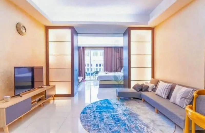 Living features: grey fabric sofa, wooden coffee table, wall-mounted TV, area rug with geometric pattern, large sliding glass door, floor-to-ceiling windows, curtains in dark brown, recessed lighting, cream painted walls, glossy tile flooring; style: modern. Bedroom features: king-size bed, bedding in light colors, nightstands, decorative wall paneling, lamps on nightstands, access to balcony, curtains in dark brown; style: modern.