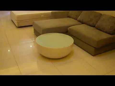 Living features: L-shape sectional sofa in grey, round coffee table with glass top, polished tile flooring, neutral wall color, soft lighting, decorative cushions, minimalist decor, large window with natural light; style: modern.