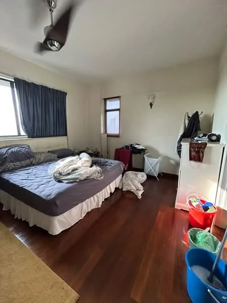 Bedroom features: queen-size bed with grey bedding, wooden flooring, floor-to-ceiling window with sheer curtains, wall-mounted light fixture, ceiling fan, white painted walls, built-in wardrobe, laundry basket, plastic storage bins, clothes on the floor.