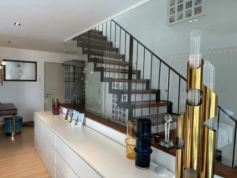 Room features: wooden staircase with black metal railing, glass partition, white cabinetry, light wooden flooring, decorative vases in blue and gold, framed artwork on wall, recessed lighting, open space layout; style: modern.