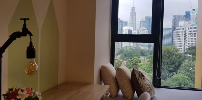 Room features: large window with city view, wooden desk, decorative lamp with exposed bulb, two decorative cushions, potted flowers, green accent wall, neutral wall colour, natural light from window; style: modern; view: city.