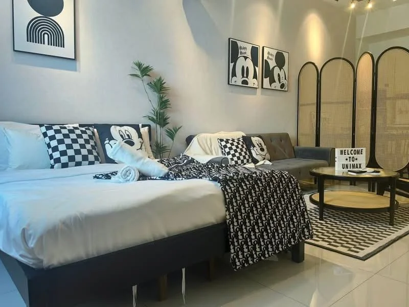 Living features: queen-size bed with black frame, white bedding with black checkered throw blanket, black and white decorative pillows, grey fabric sofa, round coffee table with black base, area rug with geometric pattern, wall-mounted artwork with abstract designs, decorative plant in pot, screen divider, recessed lighting, white painted walls, glossy tile flooring; style: modern.