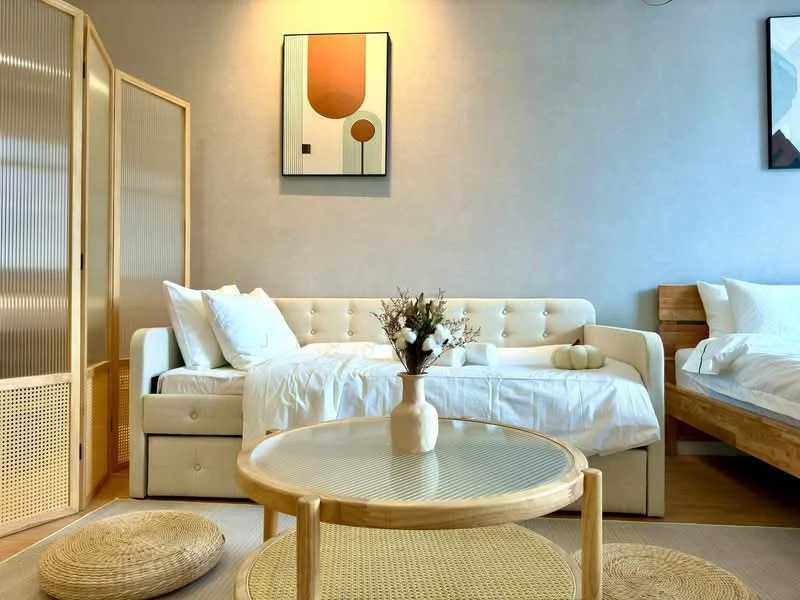 Living features: sofa bed with cream upholstery, wooden side table, round coffee table with rattan base, decorative vase with flowers, framed artwork on wall, light wood flooring, neutral wall color, woven floor cushions; style: modern.