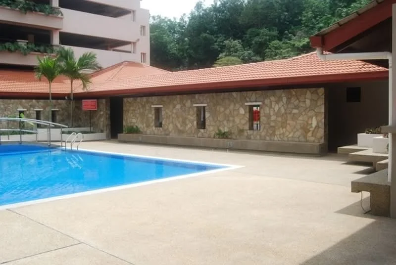 Exterior type: swimming pool deck; features: rectangular swimming pool, stone cladding on building facade, red tiled roof, paved pool deck, palm trees, surrounding greenery, multi-storey car park in background; style: tropical-modern.