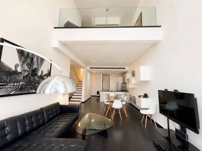 Living features: black leather sectional sofa, glass coffee table, wall-mounted TV, white dining chairs, wood dining table, high ceiling, open staircase, glass railing, dark hardwood flooring, white walls, recessed lighting, artwork on wall; style: modern. Kitchen features: white cabinetry, stainless-steel appliances, open layout, countertop, kitchen island; style: modern.