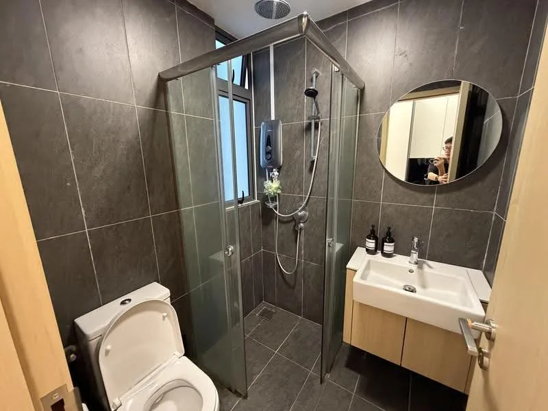 Bathroom features: walk-in shower with glass enclosure, chrome handheld shower, wall-mounted shower head, single-sink vanity with wood cabinet, round mirror, white toilet, grey tile flooring, dark grey tiled walls, soap dispenser, two black bottles on vanity; style: modern.