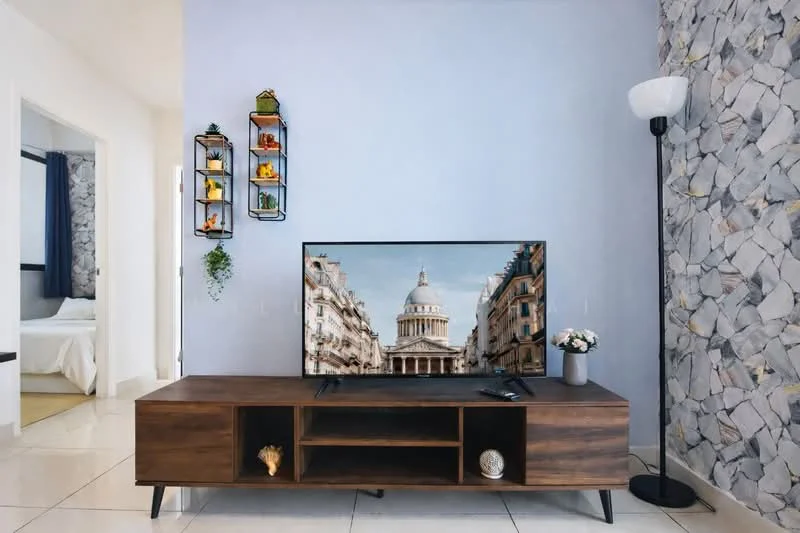 Living features: light blue painted walls, stone accent wall, white floor tiles, wooden TV console, wall-mounted TV, decorative shelving unit with plants, floor lamp, decorative items on shelves, open layout to adjacent room; style: modern.