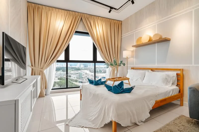 Bedroom features: queen-size bed with white bedding, wooden bed frame, two blue decorative pillows, white bedside table, table lamp with white shade, wall-mounted shelf with decorative items, floor-to-ceiling windows, sheer curtains, cream painted walls, polished tile flooring, decorative wall paneling, TV console with flat-screen TV, greenery in vase; style: modern; view: city.