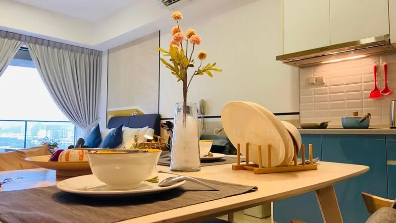 Living features: dining table with wooden base, ceramic bowl on table, cutlery on table, glass vase with flowers, white plates on wooden rack, blue cabinetry in kitchen, stainless-steel range hood, red cooking utensils on wall, floor-to-ceiling windows, curtains, grey sofa, decorative cushions; style: modern; view: city.