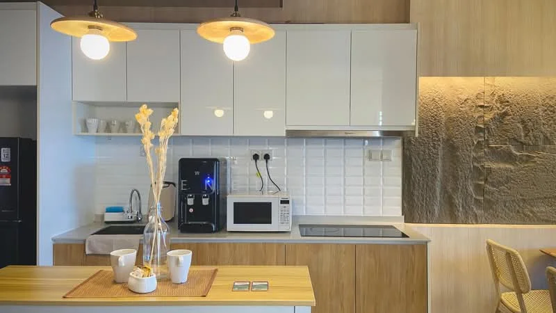 Kitchen features: white glossy cabinets, grey countertop, white subway tile backsplash, stainless-steel built-in oven, stainless-steel microwave, black coffee machine, 4-burner induction hob, kitchen island with wooden surface, vase with dried flowers, two pendant lights, ceramic cups, refrigerator, under-cabinet lighting; style: modern.