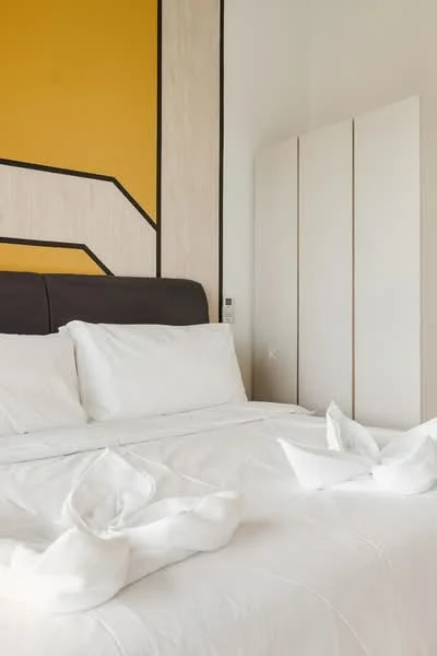 Bedroom features: king-size bed with white bedding, upholstered headboard in dark color, decorative towel arrangement on bed, yellow accent wall with geometric design, light wood paneling, built-in wardrobe with sliding doors, white bedding, nightstand with power outlet; style: modern.