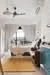 Living features: white painted walls, wooden flooring, 5-blade ceiling fan with light, large window with sheer curtains, sofa in beige fabric, coffee table, decorative wall art, area rug with geometric pattern, potted plants, study desk with chair, shelves with books, light fixture hanging from ceiling; style: modern.