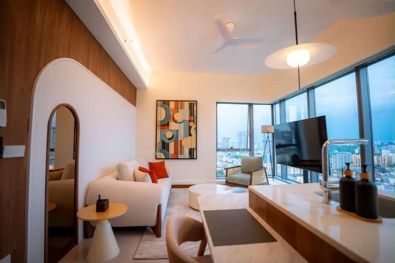 Living features: floor-to-ceiling windows, city view, 3-seater fabric sofa in white, accent pillows in orange and brown, round coffee table, area rug with geometric pattern, wall-mounted TV, wood paneling on wall, decorative artwork on wall, ceiling fan with light, recessed lighting, open-plan layout with kitchen, bar stools at kitchen counter; style: modern.