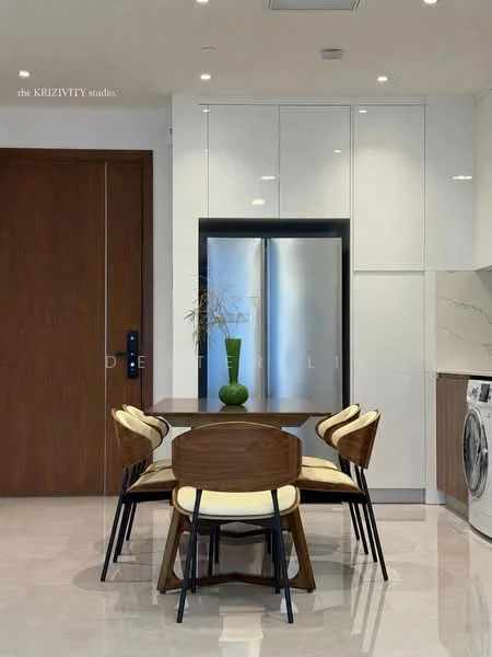 Kitchen features: white cabinetry with glossy finish, stainless-steel fridge, wooden dining table, 4 wooden dining chairs with cream upholstery, white marble countertop, built-in washing machine, recessed ceiling lights, large wall-mounted mirror, neutral tile flooring, green decorative vase on table; style: modern.