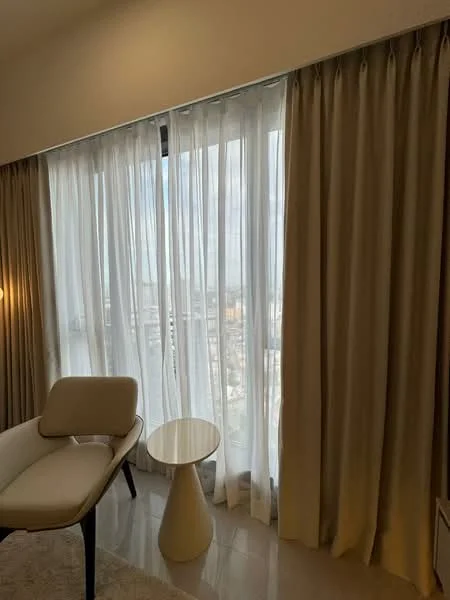 Room features: floor-to-ceiling windows, sheer curtains, curtains in beige, light-coloured wall, cream wall, white ceiling, marble flooring, small round side table, upholstered chair in light colour, area rug; style: modern; view: city.