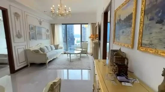 Living features: 3-seater fabric sofa in light grey, glass coffee table, area rug with geometric pattern, chandelier with multiple bulbs, floor-to-ceiling windows, curtains in light fabric, decorative wall paneling, framed artwork on walls, console table with decorative items, wall-mounted TV, wooden flooring with glossy finish; style: luxury; view: city.