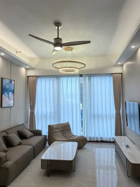 Living features: 3-seater fabric sofa in beige, accent chair in brown, round coffee table with white surface, TV console with white finish, wall-mounted TV, chandelier with circular design, 5-blade ceiling fan with light, floor-to-ceiling windows with sheer curtains, light-colored wall with wood paneling, polished tile flooring, area rug with circular pattern; style: modern; view: city.