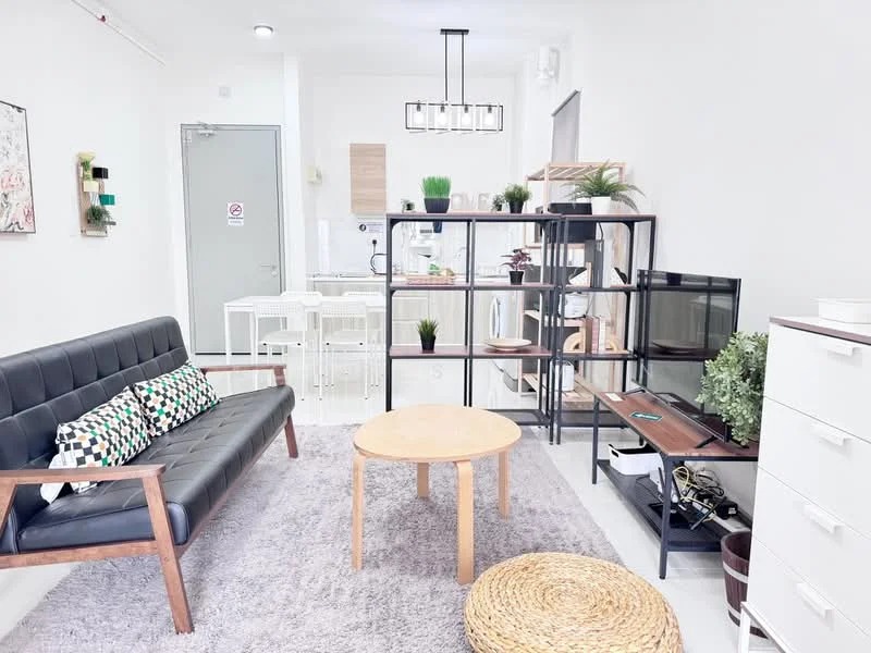 Living features: black leather sofa with wooden frame, decorative cushions with geometric patterns, round wooden coffee table, area rug with neutral color, TV console with storage, wall-mounted TV, open shelving unit with plants, white walls, recessed ceiling lights, large window with natural light; style: modern. Dining features: white dining table for four, white dining chairs, open kitchen layout, wooden shelving unit, kitchen appliances visible in background; style: modern.