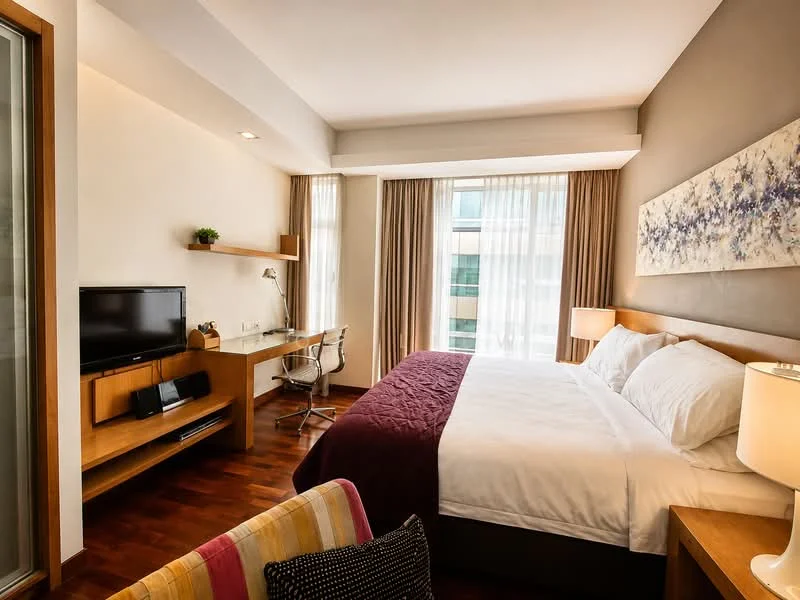 Bedroom features: king-size bed with white bedding, upholstered headboard, wooden bedside tables, table lamps with fabric shades, wall-mounted TV, wooden desk with chair, large window with sheer curtains, wooden flooring, decorative artwork on wall, built-in wardrobe; style: modern; view: city.