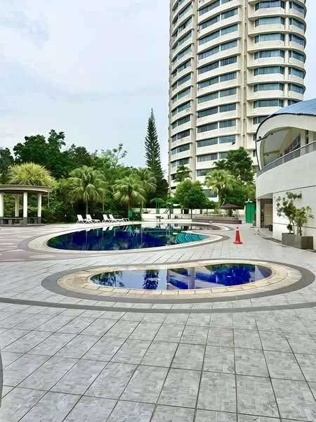 Exterior type: high-rise condominium; features: infinity pool, pool deck with loungers, landscaped garden with palm trees, tiled flooring around pool area, surrounding greenery, modern architectural design, multiple levels of the building, outdoor seating area, safety cone visible; style: modern.