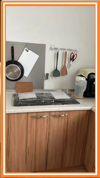 Kitchen features: laminate cabinetry in wood finish, white countertop, built-in cooktop, kitchen utensils hanging on wall, black frying pan, white cutting board, grey wall panel, coffee machine, spatula, scissors, wooden spoon, cork trivet; style: modern.