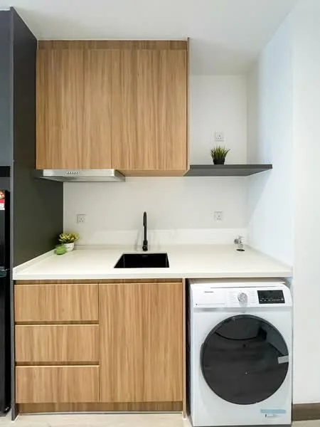 Laundry features: wood cabinetry with natural finish, white countertop, single-bowl black sink, black gooseneck faucet, built-in washing machine, open shelving above countertop, refrigerator, decorative plant on shelf; style: modern.