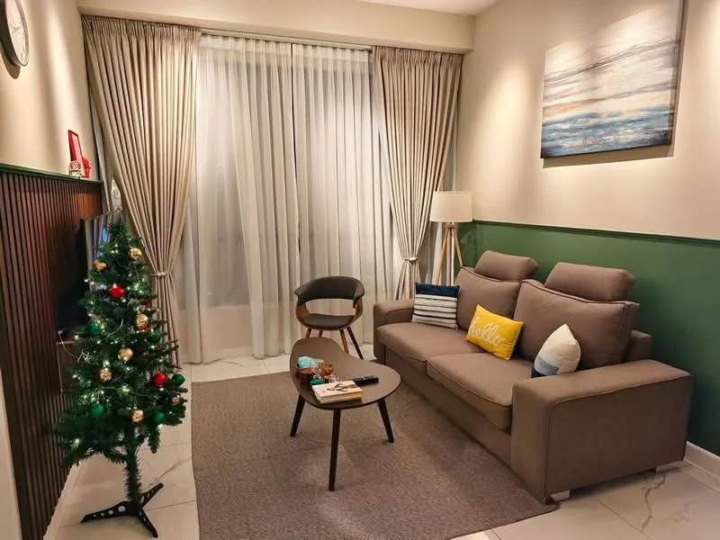 Living features: 3-seater fabric sofa in grey, decorative cushions in various colors, wooden coffee table, area rug in neutral color, floor-to-ceiling windows with sheer curtains, green accent wall, wall-mounted clock, framed artwork on wall, decorative Christmas tree, table lamp, wooden chair; style: modern.