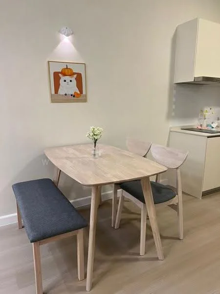 Dining features: light wood dining table, grey upholstered bench, two wooden dining chairs, decorative wall art with a cat, small vase with flowers, light fixture on wall, neutral wall color, laminate flooring; style: modern. Kitchen features: white cabinetry, countertop with kitchen items visible, neutral wall color; style: modern.