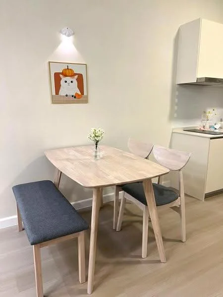 Dining features: light wood dining table, grey upholstered bench, two wooden dining chairs, framed artwork on wall, small vase with flowers, light fixture on wall, light wood flooring, neutral painted walls; style: modern. Kitchen features: white cabinetry, countertop, kitchen appliances visible; style: modern.