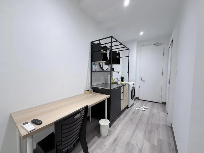 Room features: white painted walls, light wood laminate flooring, desk with light wood surface, black office chair, built-in shelving unit, washing machine, kitchenette with sink, refrigerator, door to hallway, ceiling lights; style: modern.