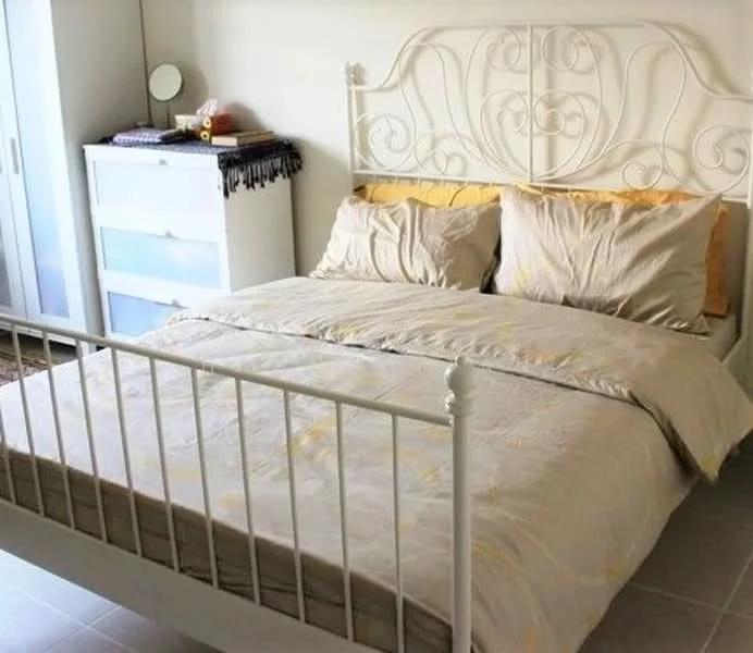 Bedroom features: queen-size bed with white metal frame, beige bedding with decorative pillows, white wardrobe with sliding doors, chest of drawers with white finish, decorative items on top of wardrobe, light-colored walls, tile flooring, window with natural light; style: modern.