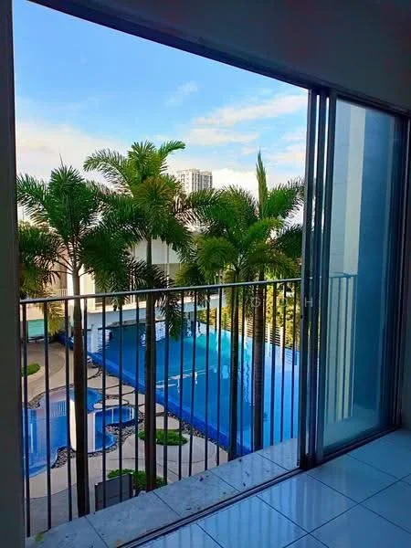 Room features: floor-to-ceiling sliding glass door, balcony with railing, view of swimming pool, view of palm trees, white tiled flooring, natural light from large window, blue sky visible, outdoor space visible; style: modern; view: pool.